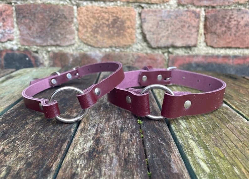 May include: Two burgundy leather collars with silver metal rings. The collars are lying on a wooden surface.