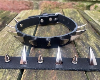 May include: A black leather collar with silver-colored metal spikes. The collar has a buckle closure and is displayed with a matching strip of leather with additional spikes. The spikes are various shapes and sizes.
