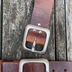 May include: A brown leather belt featuring a textured, distressed surface and a large, square-shaped brushed silver metal buckle. The belt strap is secured to the buckle with three small silver-toned metal rivets.