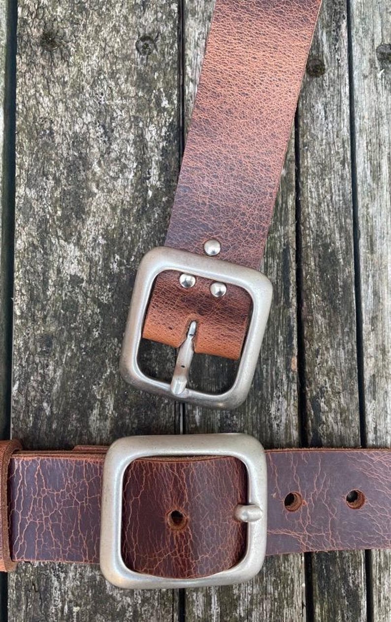 May include: A brown leather belt featuring a textured, distressed surface and a large, square-shaped brushed silver metal buckle. The belt strap is secured to the buckle with three small silver-toned metal rivets.