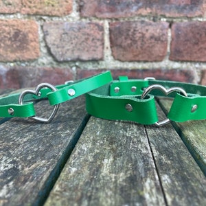May include: Two green leather collars with silver heart-shaped buckles. The collars are lying on a wooden surface.
