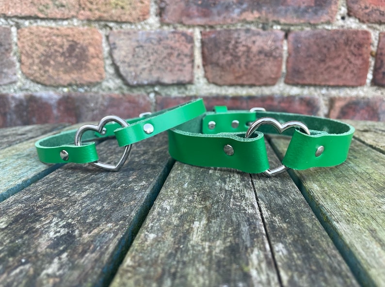 May include: Two green leather collars with silver heart-shaped buckles. The collars are lying on a wooden surface.