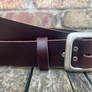 May include: A dark brown leather belt featuring a rectangular silver-toned metal buckle. The belt includes a matching leather keeper loop and two silver-toned metal rivets securing the buckle to the strap, showcasing a smooth finish.