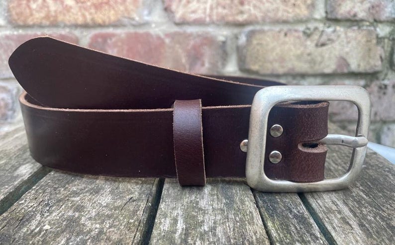 May include: A dark brown leather belt featuring a rectangular silver-toned metal buckle. The belt includes a matching leather keeper loop and two silver-toned metal rivets securing the buckle to the strap, showcasing a smooth finish.