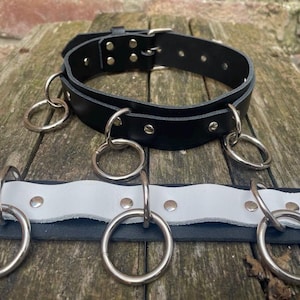 May include: A black leather collar featuring a silver-toned buckle and multiple silver metal O-rings attached with rivets. Below it lies a flat black and white leather strap also adorned with evenly spaced silver metal O-rings and rivets.