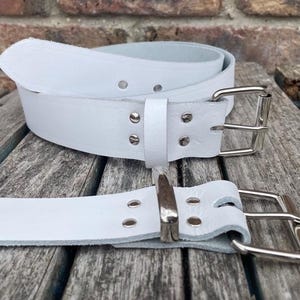 May include: Two white leather belts with silver buckles are shown on a wooden surface. The belts are rolled and laid out, showcasing their design and hardware. The belts are a classic accessory.