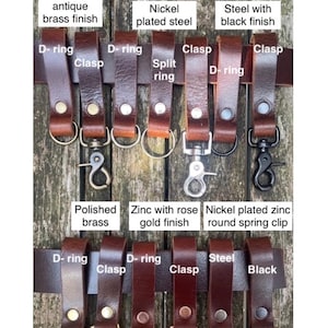 May include: A collection of oak-coloured buffalo leather belt loops, each secured with a single large rivet. The loops feature various metal hardware attachments including D-rings, clasps, split rings, and spring clips in antique brass, nickel, black, and rose gold finishes.