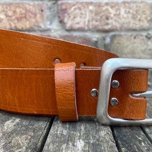 May include: A tan leather belt featuring a smooth, matte finish and a rectangular silver-toned metal buckle. The belt includes a matching leather keeper loop and is secured with two visible silver-toned metal rivets near the buckle.