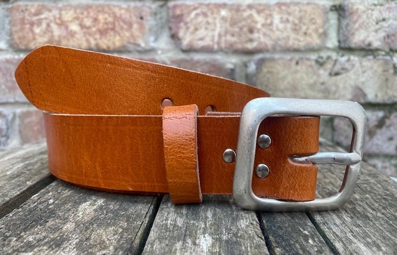 May include: A tan leather belt featuring a smooth, matte finish and a rectangular silver-toned metal buckle. The belt includes a matching leather keeper loop and is secured with two visible silver-toned metal rivets near the buckle.