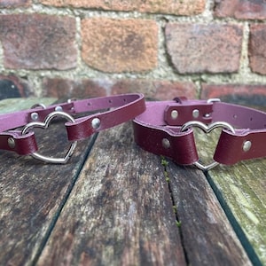 May include: Two burgundy leather chokers with silver heart-shaped clasps. The chokers are lying on a wooden surface.