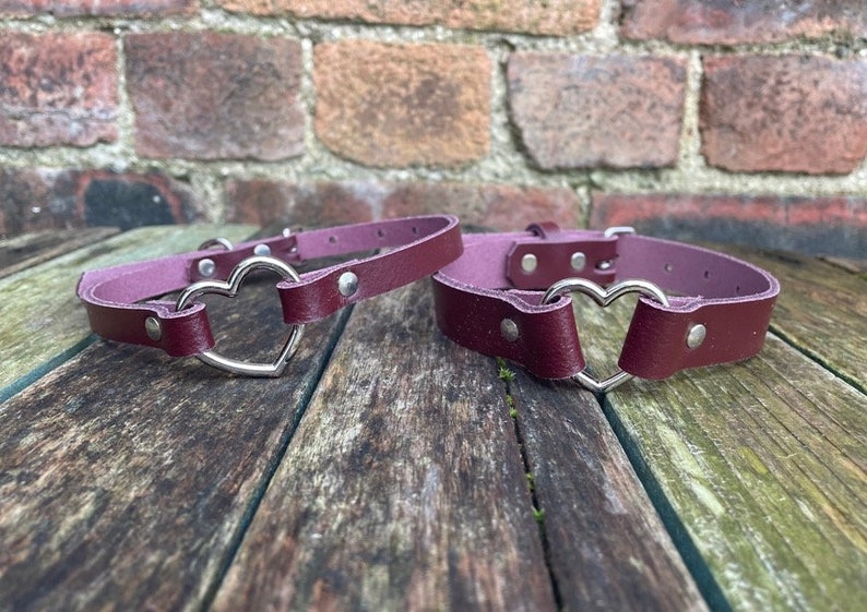 May include: Two burgundy leather chokers with silver heart-shaped clasps. The chokers are lying on a wooden surface.