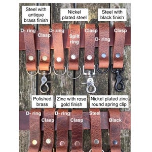 May include: A collection of tan buffalo leather belt loops featuring various metal hardware attachments including D-rings, clasps, split rings, and spring clips. The hardware finishes include antique brass, nickel, black, polished brass, and rose gold.