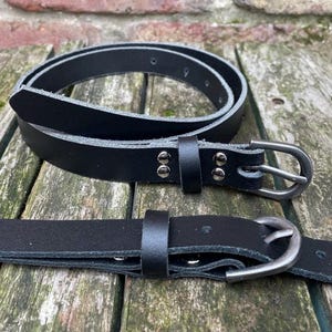 Peut inclure: Deux ceintures en cuir noir avec des surfaces lisses et une finition mate. Chaque ceinture présente une boucle en métal simple, un passant en cuir assorti et deux rivets en métal argenté fixant l'extrémité de la boucle. Les ceintures sont fines.