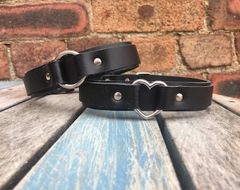 Puede incluir: Dos collares de cuero negro con herrajes de metal plateado. Un collar incluye una anilla circular, mientras que el otro presenta una anilla en forma de corazón. Ambas bandas están aseguradas con remaches de metal plateado en las correas.