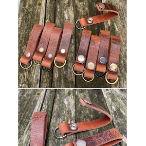 May include: A collection of brown leather belt loops featuring metal D-rings. Some loops are permanently closed with rivets, while others include metal press studs for opening and closing. The hardware comes in various metallic finishes.