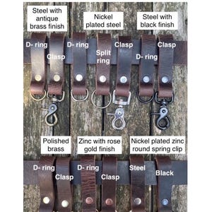 May include: A collection of dark brown buffalo leather belt loops featuring various metal hardware attachments including D-rings, clasps, split rings, and spring clips. The hardware finishes include antique brass, nickel, black, polished brass, and rose gold.