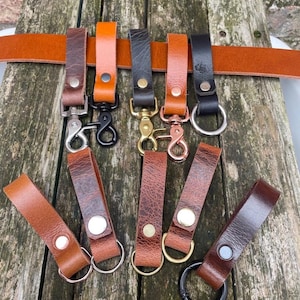 May include: A collection of leather belt loops and key keepers in various shades of brown and black, featuring metal rivets and hardware including silver, gold, copper, and black swivel clips and circular rings attached to a long leather belt.