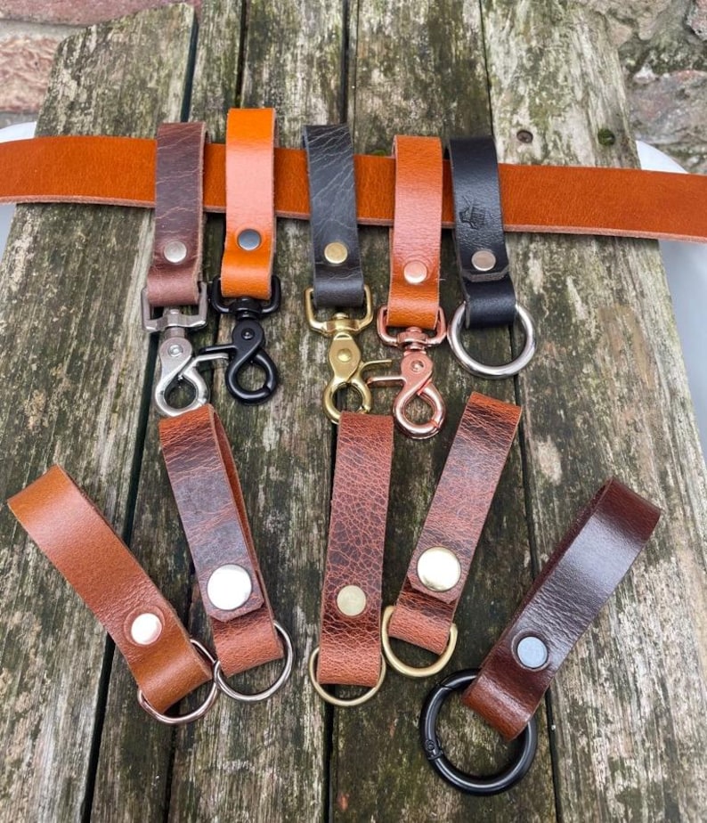 May include: A collection of leather belt loops and key keepers in various shades of brown and black, featuring metal rivets and hardware including silver, gold, copper, and black swivel clips and circular rings attached to a long leather belt.