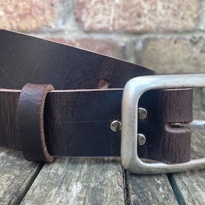 May include: A dark brown leather belt featuring a wide, rectangular silver-toned metal buckle. The belt includes a matching leather keeper loop and two silver-toned metal rivets securing the buckle to the strap, showing a textured finish.