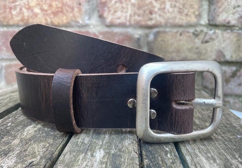 May include: A dark brown leather belt featuring a wide, rectangular silver-toned metal buckle. The belt includes a matching leather keeper loop and two silver-toned metal rivets securing the buckle to the strap, showing a textured finish.