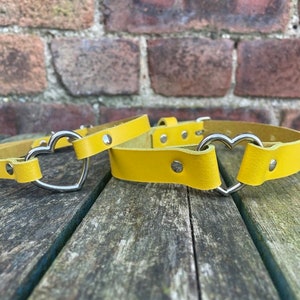 May include: Two yellow leather belts with heart-shaped metal buckles. The belts are lying on a wooden surface.