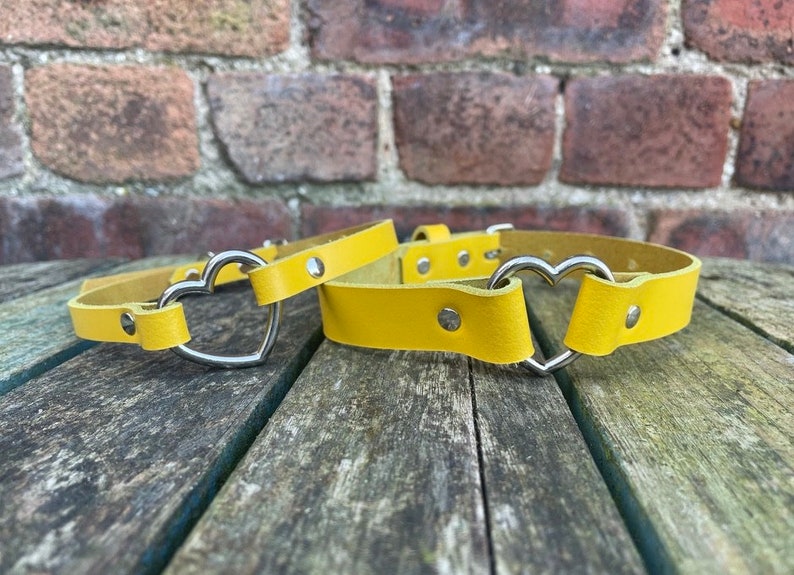May include: Two yellow leather belts with heart-shaped metal buckles. The belts are lying on a wooden surface.