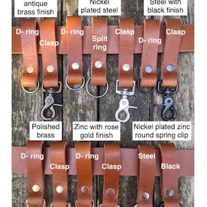 May include: A collection of brandy-coloured buffalo leather belt loops, each secured with a single large rivet. The loops feature various metal hardware attachments including D-rings, split rings, clasps, and spring clips in brass, nickel, steel, and rose gold finishes.