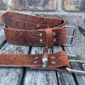 May include: Two brown leather belts with a textured, reptile-like pattern. The belts have silver-coloured metal buckles and rivets. One belt is coiled, and the other is laid out flat. The belts are on a weathered wooden surface.