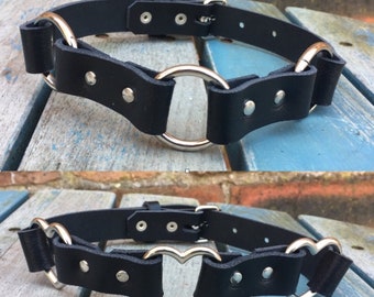 Puede incluir: Dos collares de cuero negro con herrajes de metal plateado. El collar superior tiene un anillo redondo grande, mientras que el collar inferior tiene dos anillos en forma de corazón.