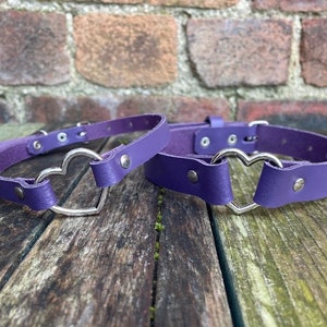 May include: Two purple leather chokers with silver heart-shaped buckles. The chokers are on a wooden surface.