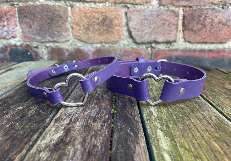 May include: Two purple leather chokers with silver heart-shaped buckles. The chokers are on a wooden surface.