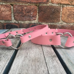 May include: Two pink leather choker necklaces with silver heart-shaped buckles. The necklaces are on a wooden surface.