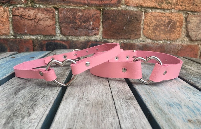 May include: Two pink leather choker necklaces with silver heart-shaped buckles. The necklaces are on a wooden surface.