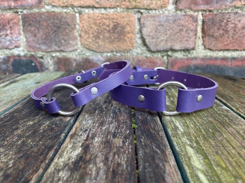 May include: Two purple leather collars with silver metal rings. The collars are on a wooden surface.