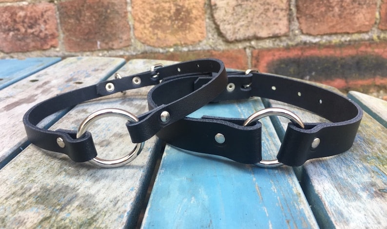 May include: Two black leather collars with silver metal rings. The collars are on a blue wooden surface.