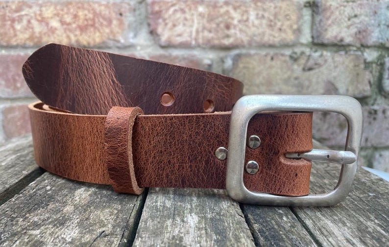 May include: A brown leather belt featuring a textured surface with visible grain patterns, a single leather keeper loop, and a rectangular brushed silver-tone metal buckle secured by two metal rivets on the strap.