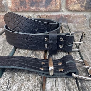 Può includere: Due cinture in pelle nera con una texture a pelle di coccodrillo sono esposte su una superficie di legno. Le cinture hanno fibbie e rivetti color argento. Una è arrotolata, l'altra è piatta, evidenziando la texture e il design dettagliati.