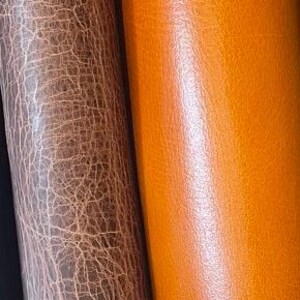 May include: A collection of six vertical leather rolls displayed side by side. The textures vary from smooth, solid dark brown and vibrant orange to distressed, cracked surfaces in shades of light brown, dark brown, and charcoal gray.