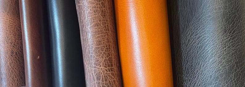 May include: A collection of six vertical leather rolls displayed side by side. The textures vary from smooth, solid dark brown and vibrant orange to distressed, cracked surfaces in shades of light brown, dark brown, and charcoal gray.