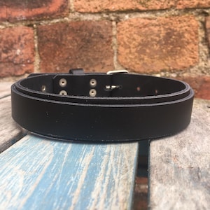 Puede incluir: Un cinturón de cuero negro con una hebilla plateada y múltiples filas de costuras. El cinturón tiene cinco filas de agujeros para ajustar el ajuste.