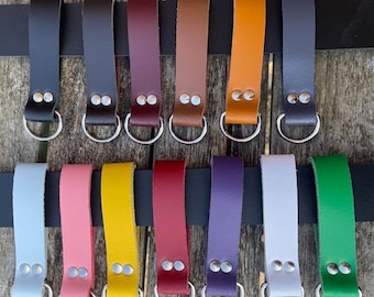 May include: A selection of leather key fobs in diverse colours, such as black, brown, orange, and green. Each fob features a metal ring and is attached to a black strap. The fobs are presented against a wooden backdrop.