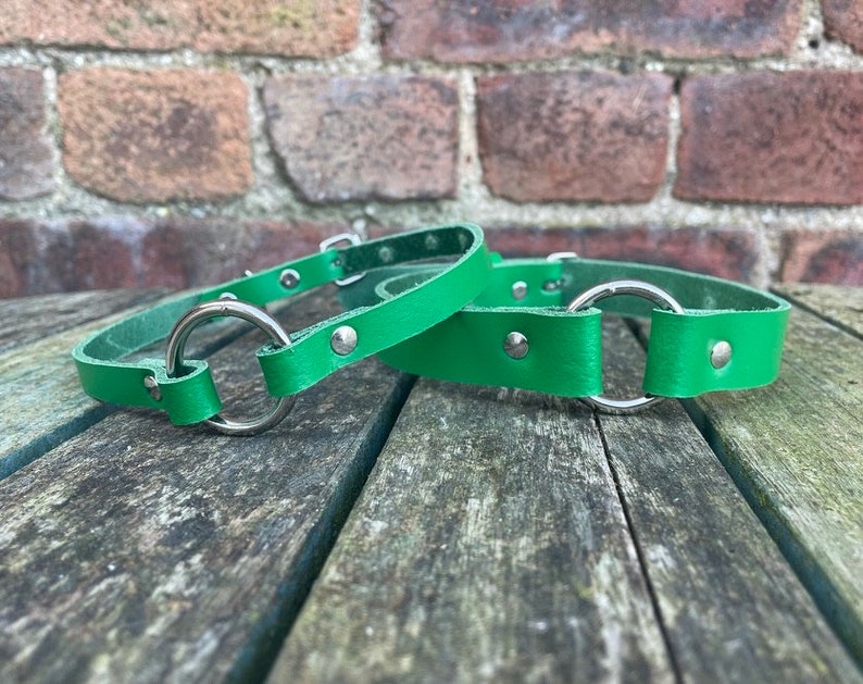 May include: Two green leather collars with silver metal rings and studs. The collars are on a wooden surface.