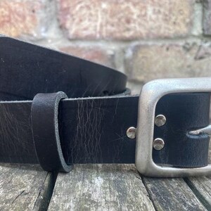 May include: A black leather belt featuring a smooth, matte finish and a rectangular silver-toned metal buckle. The belt includes a matching leather keeper loop and two silver-toned metal rivets securing the buckle to the strap.