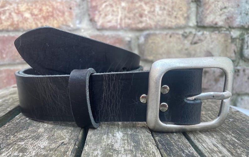 May include: A black leather belt featuring a smooth, matte finish and a rectangular silver-toned metal buckle. The belt includes a matching leather keeper loop and two silver-toned metal rivets securing the buckle to the strap.