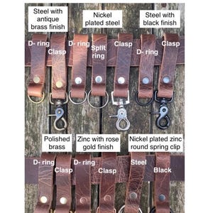 May include: A collection of light brown buffalo leather belt loops featuring various metal hardware attachments including D-rings, split rings, and clasps in antique brass, nickel-plated steel, black steel, polished brass, and rose gold finishes.