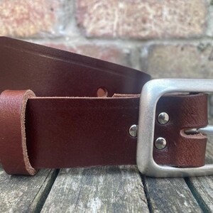 May include: A dark brown leather belt featuring a smooth finish, a single leather keeper loop, and a rectangular silver-toned metal buckle secured with two metal rivets. The belt strap has a pointed end and visible stitching details.