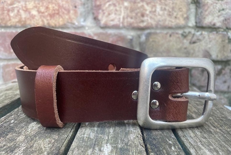 May include: A dark brown leather belt featuring a smooth finish, a single leather keeper loop, and a rectangular silver-toned metal buckle secured with two metal rivets. The belt strap has a pointed end and visible stitching details.