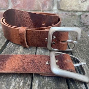 May include: Two brown leather belts with a visible grain texture and a distressed finish. Each belt features a large, square-shaped brushed silver metal buckle secured with two rivets, and a matching leather keeper loop on the coiled belt.