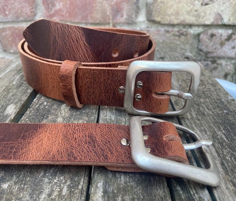 May include: Two brown leather belts with a visible grain texture and a distressed finish. Each belt features a large, square-shaped brushed silver metal buckle secured with two rivets, and a matching leather keeper loop on the coiled belt.