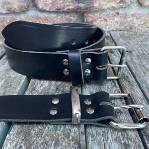 May include: Two black leather belts with silver-colored metal buckles. One belt is coiled, and the other is laid out flat. The belts are on a weathered wooden surface, with a brick wall in the background.